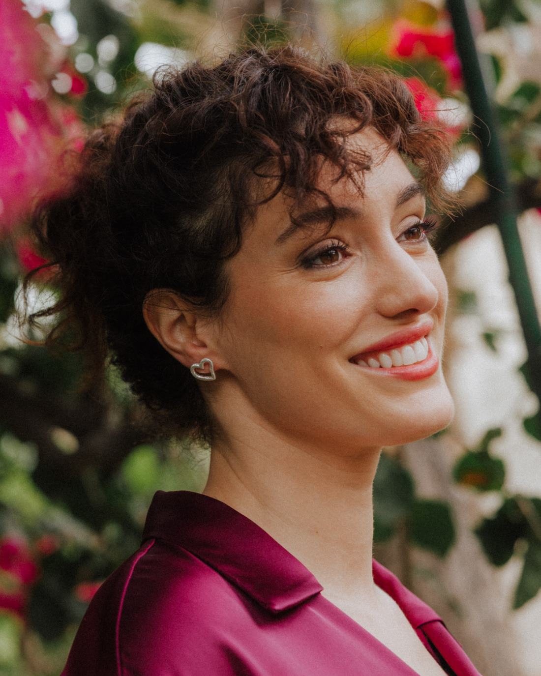 Primer plano de una persona sonriendo, llevando un pendiente plateado con forma de corazón irregular y diseño abierto ajustado al lóbulo. El fondo muestra vegetación y flores desenfocadas en tonos rosas y verdes.
