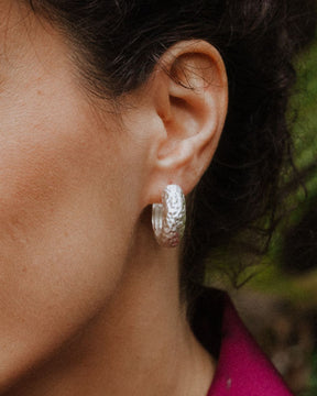 Primer plano de un aro plateado con textura martillada colocado en la oreja de una persona, con fondo natural desenfocado.