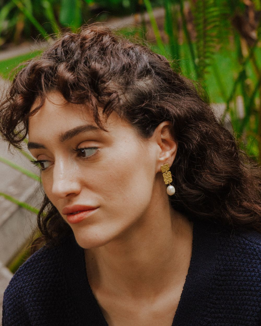 Pendiente dorado con dos piezas rectangulares texturizadas y una perla colgante, lucido por una modelo en un entorno natural con vegetación al fondo. La joya destaca sutilmente sobre un jersey oscuro.