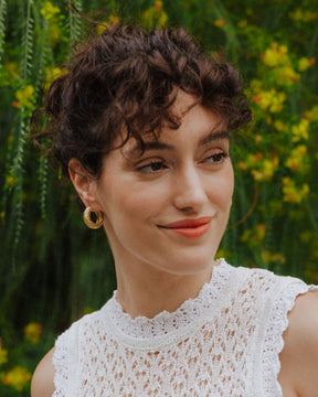 Pendientes dorados de aro grueso con textura martillada lucidos por una modelo, que lleva un top blanco de encaje. El fondo muestra un entorno natural con vegetación verde y flores amarilla.