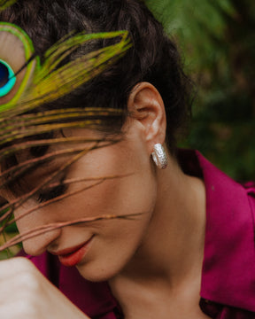 Primer plano de la modelo con labios rojos y blusa color magenta inclinando la cabeza hacia abajo. Lleva aros plateados de textura martillada y sostiene una pluma de pavo real frente a su rostro, con fondo natural desenfocado.