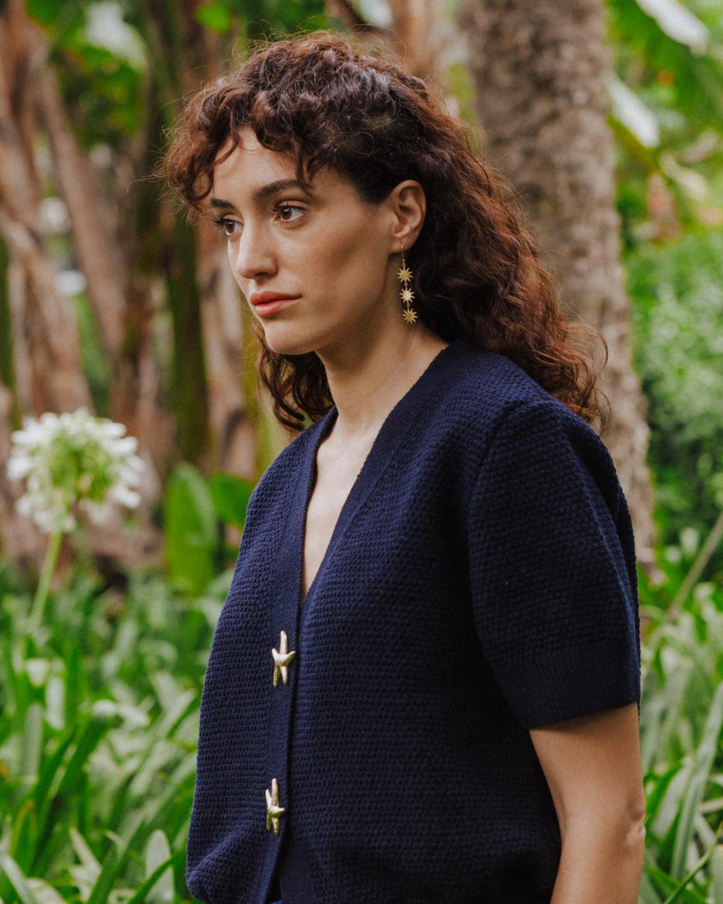 Persona de pie en un entorno natural, llevando un pendiente largo dorado formado por tres pequeñas piezas en forma de sol de bordes irregulares y rayos finos, unidas entre sí con pequeñas argollas. El pendiente cuelga desde un gancho fino. La persona viste una chaqueta de punto azul marino y aparece rodeada de vegetación y troncos de árboles desenfocados al fondo.