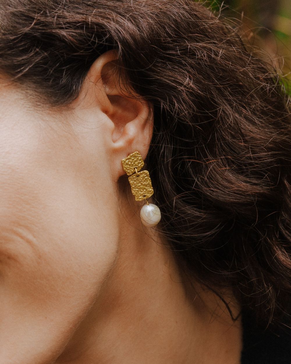 Pendiente dorado formado por dos rectángulos texturizados unidos, del que cuelga una perla ovalada. La pieza se muestra puesta en la oreja de una modelo, con un fondo natural y tonos verdes suaves desenfocados.