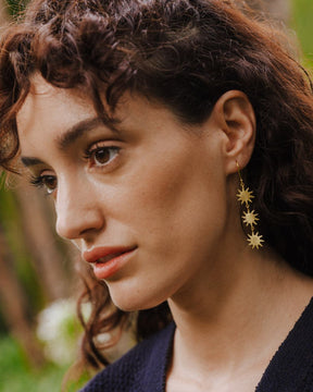 Primer plano de una persona con el pelo rizado llevando un pendiente largo dorado formado por tres pequeñas piezas en forma de sol de bordes irregulares y rayos finos, unidas entre sí con pequeñas argollas. El pendiente cuelga desde un gancho fino y queda sobre el cuello