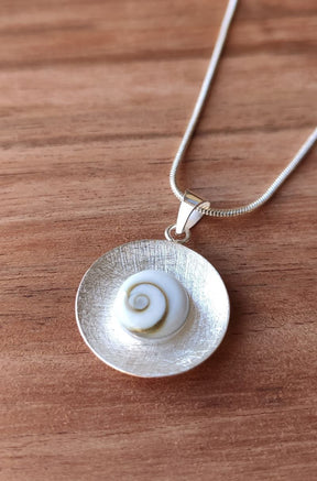 Primer plano del colgante que presenta una piedra de Santa Lucía enmarcada en un disco de textura cepillada con collar bañado en plata 925 encima de un fondo de madera