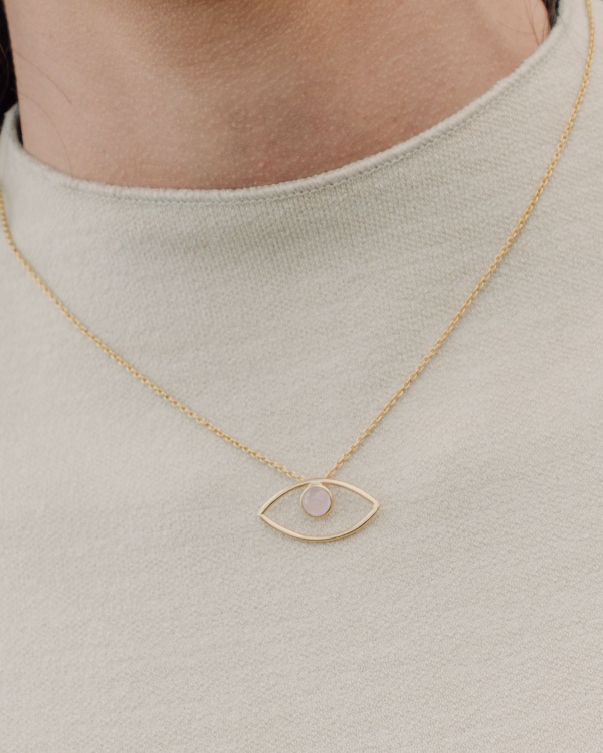 Primer plano del cuello de la modelo con un jersey blanco y el collar bañado en oro de 18 quilates con un colgante en forma de ojo y piedra semipreciosa en Cuarzo Rosa de Isla Vurma