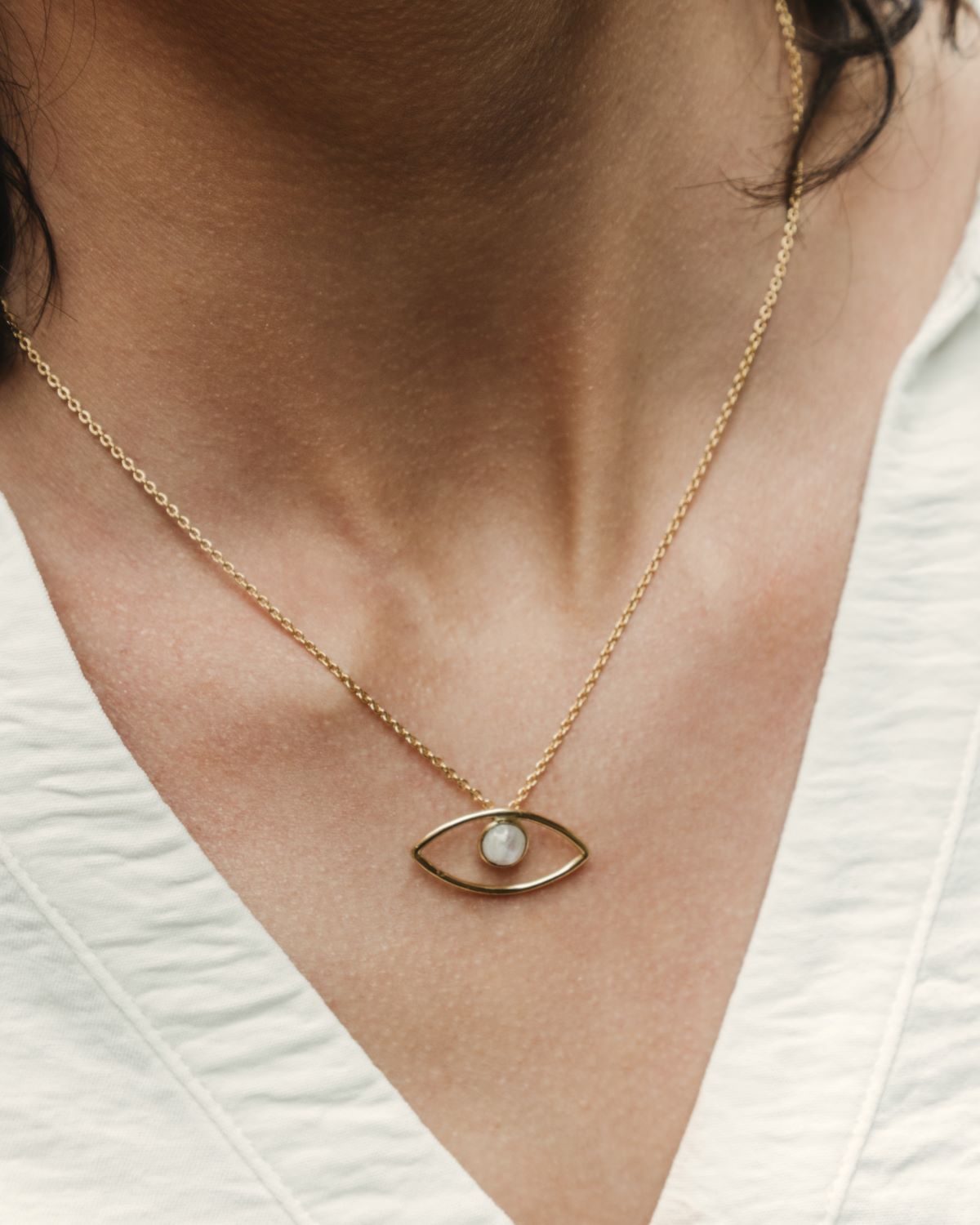 Primer plano del cuello de la modelo con una camiseta blanca y el collar bañado en oro de 18 quilates con un colgante en forma de ojo y piedra semipreciosa en Piedra de Luna de Isla Vurma