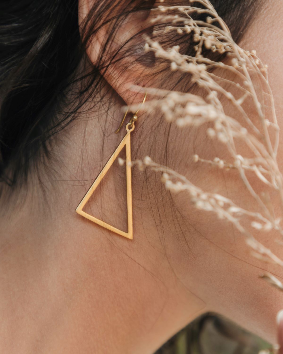 Pendiente triangular grande de latón bañado en oro 18k lucido en la oreja, con ramas finas desenfocadas en primer plano.