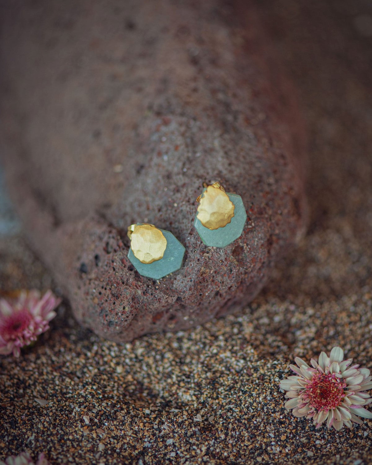 Pendientes dorados con base facetada en Aqua Calcedonia situados sobre una roca oscura, acompañados de pequeñas flores secas alrededor.