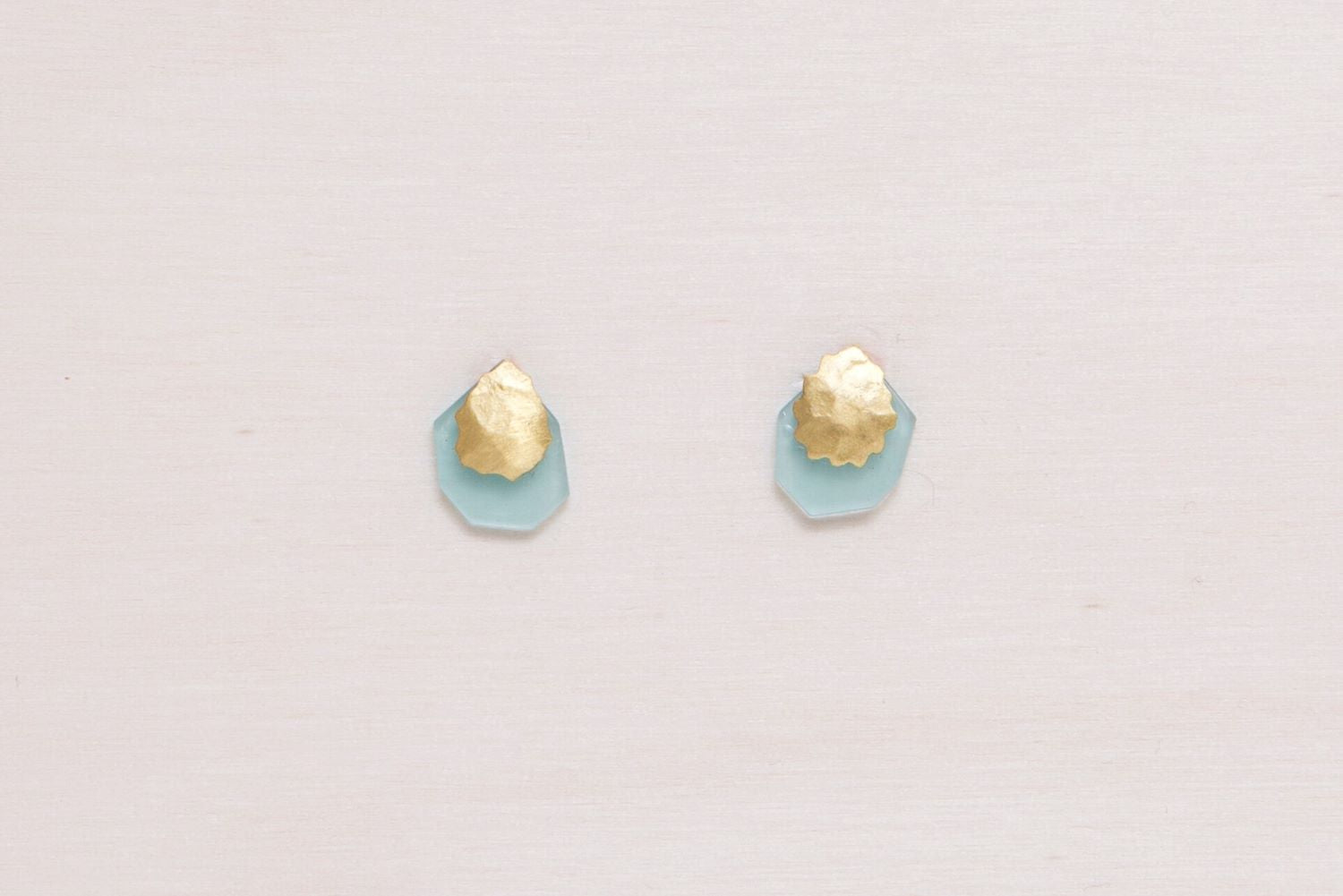 Pendientes de botón en latón bañado en oro 18k con pieza martillada orgánica y base geométrica en piedra semipreciosa Aqua Calcedonia, colocados sobre una roca de textura granulada.
