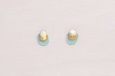 Pendientes de botón en latón bañado en oro 18k con pieza martillada orgánica y base geométrica en piedra semipreciosa Aqua Calcedonia, colocados sobre una roca de textura granulada.