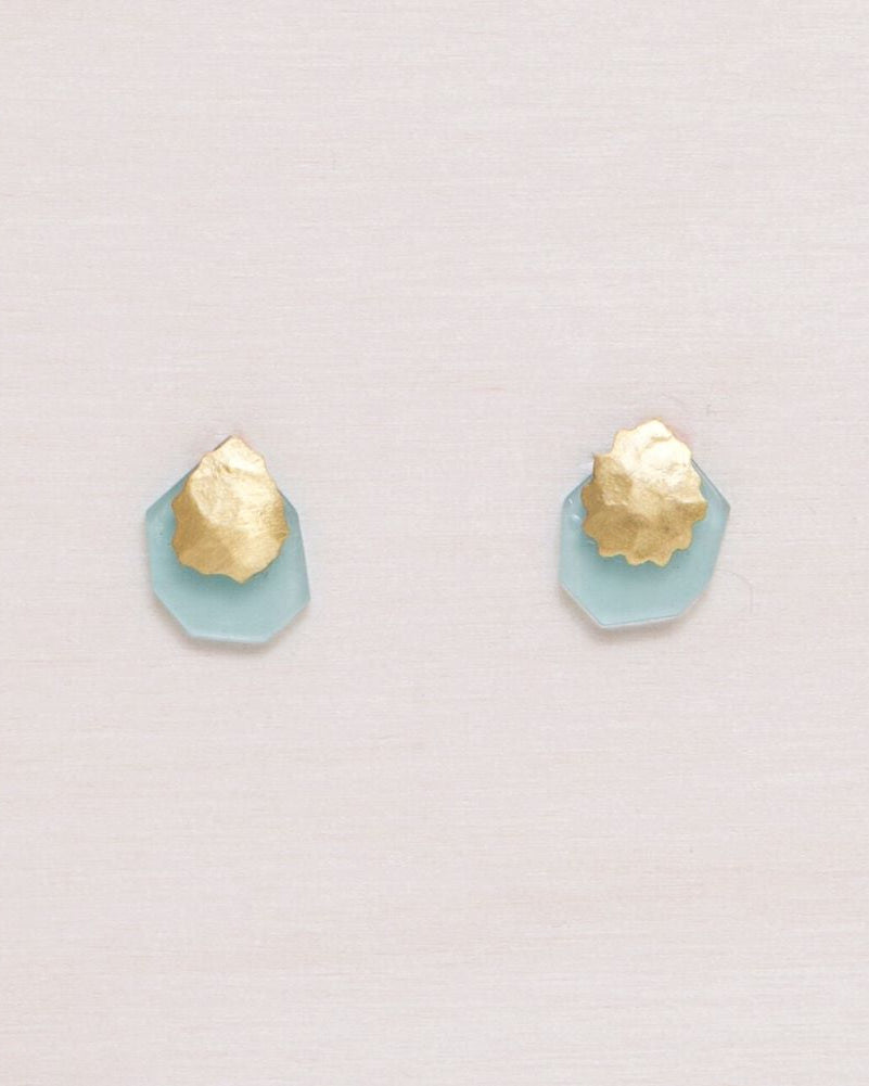 Pendientes de botón en latón bañado en oro 18k con pieza martillada orgánica y base geométrica en piedra semipreciosa Aqua Calcedonia, colocados sobre una roca de textura granulada.