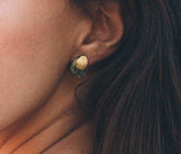 Pendiente pequeño con base irregular bañada en oro y piedra facetada de labradorita, colocado en el lóbulo de la oreja de una modelo con el cabello castaño suelto.