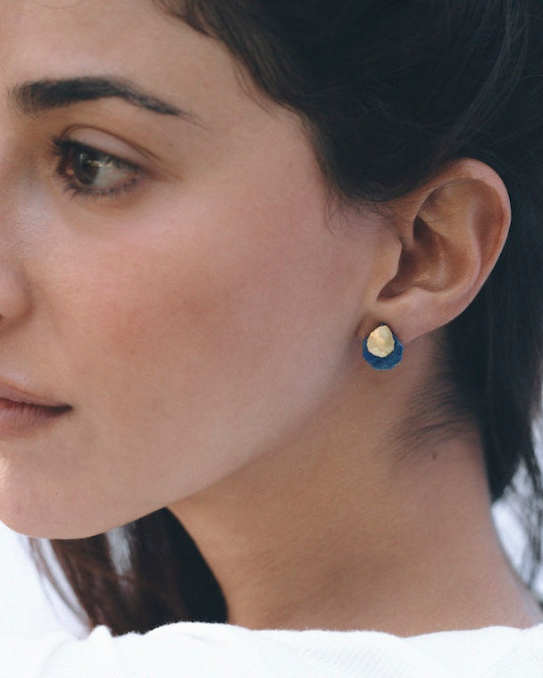 Pendientes de botón con una pieza facetada de latón bañado en oro en la parte superior y una base de piedra semipreciosa lapislázuli, colocados en la oreja de una modelo de perfil.