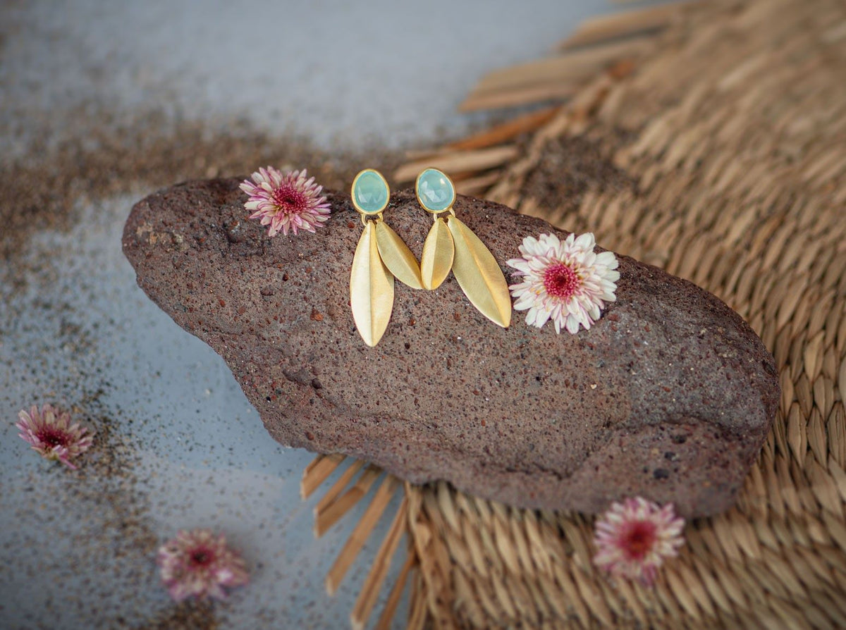 Pareja de pendientes dorados colocados sobre una piedra volcánica, cada pendiente tiene una pieza ovalada en tono Aqua Calcedonia en la parte superior y hojas metálicas doradas colgando, alrededor de la piedra hay pequeñas flores secas en tonos rosados y el fondo combina arena fina y una superficie tejida de fibras naturales
