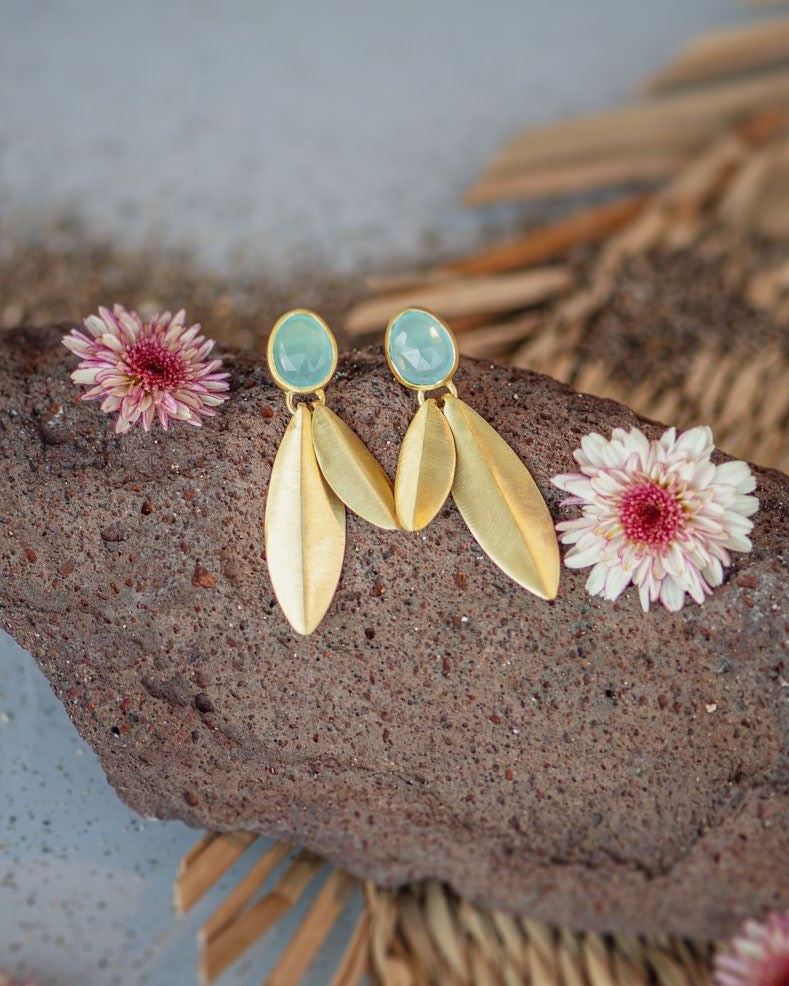 Pareja de pendientes dorados colocados sobre una piedra volcánica, cada pendiente tiene una pieza ovalada en tono Aqua Calcedonia en la parte superior y hojas metálicas doradas colgando, alrededor de la piedra hay pequeñas flores secas en tonos rosados y el fondo combina arena fina y una superficie tejida de fibras naturales