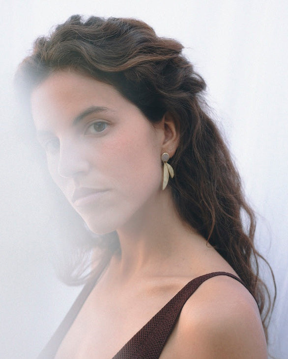 Pendientes dorados con diseño de hojas y Piedra de Luna combinados con un vestido de tirantes, mostrados en un retrato vertical con efecto bruma en primer plano.