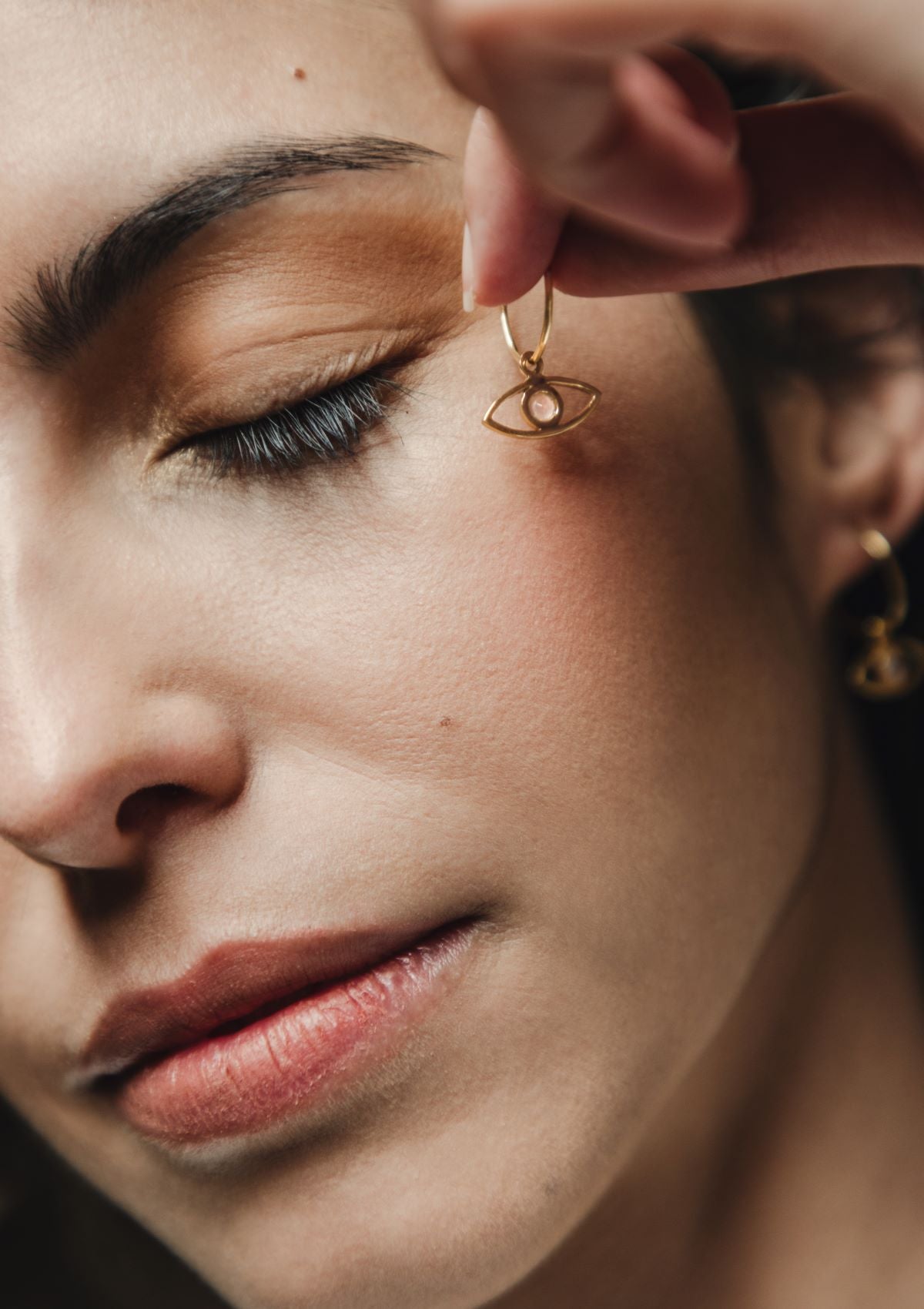 Primer plano de la modelo sosteniendo con la mano los pendientes en forma de ojo bañados en oro de 18k y con piedra semipreciosa en Rosa Calcedonia de Isla Vurma