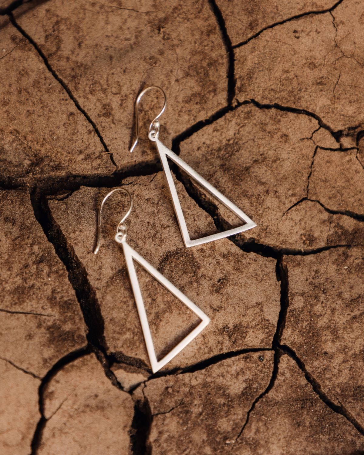 Pendientes triangulares grandes en latón bañado en plata 925 apoyados sobre una superficie seca y agrietada de tono tierra.