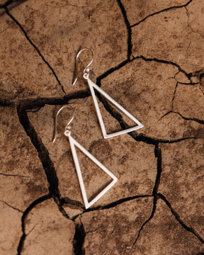 Pendientes triangulares grandes en latón bañado en plata 925 apoyados sobre una superficie seca y agrietada de tono tierra.