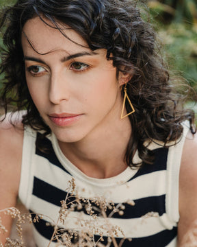 Pendiente triangular dorado de latón bañado en oro 18k en la oreja de una modelo de pelo rizado, fotografiada en un entorno natural.