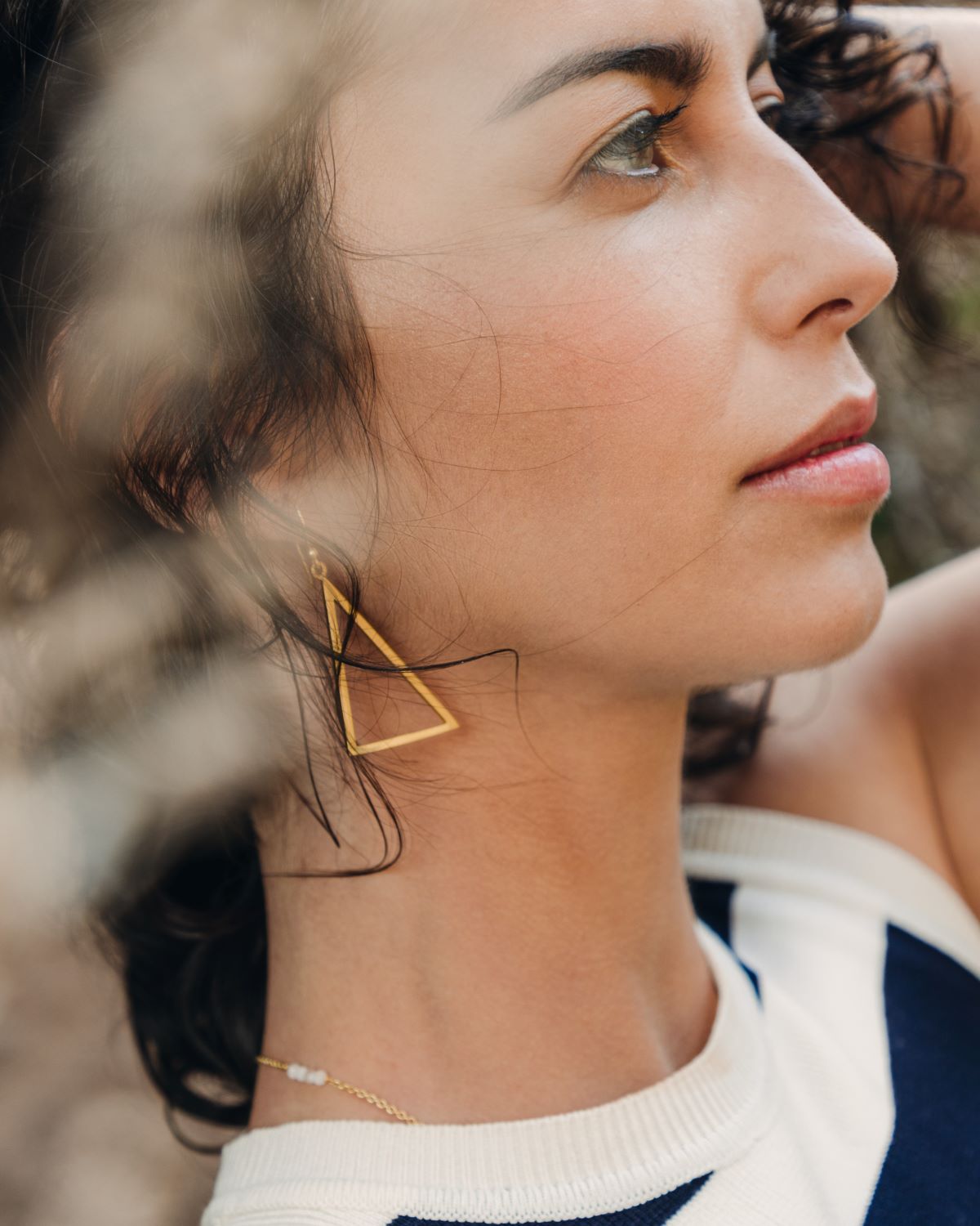 Pendiente triangular de latón bañado en oro 18k llevado por una modelo morena con luz suave y fondo natural.