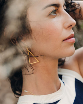 Pendiente triangular de latón bañado en oro 18k llevado por una modelo morena con luz suave y fondo natural.