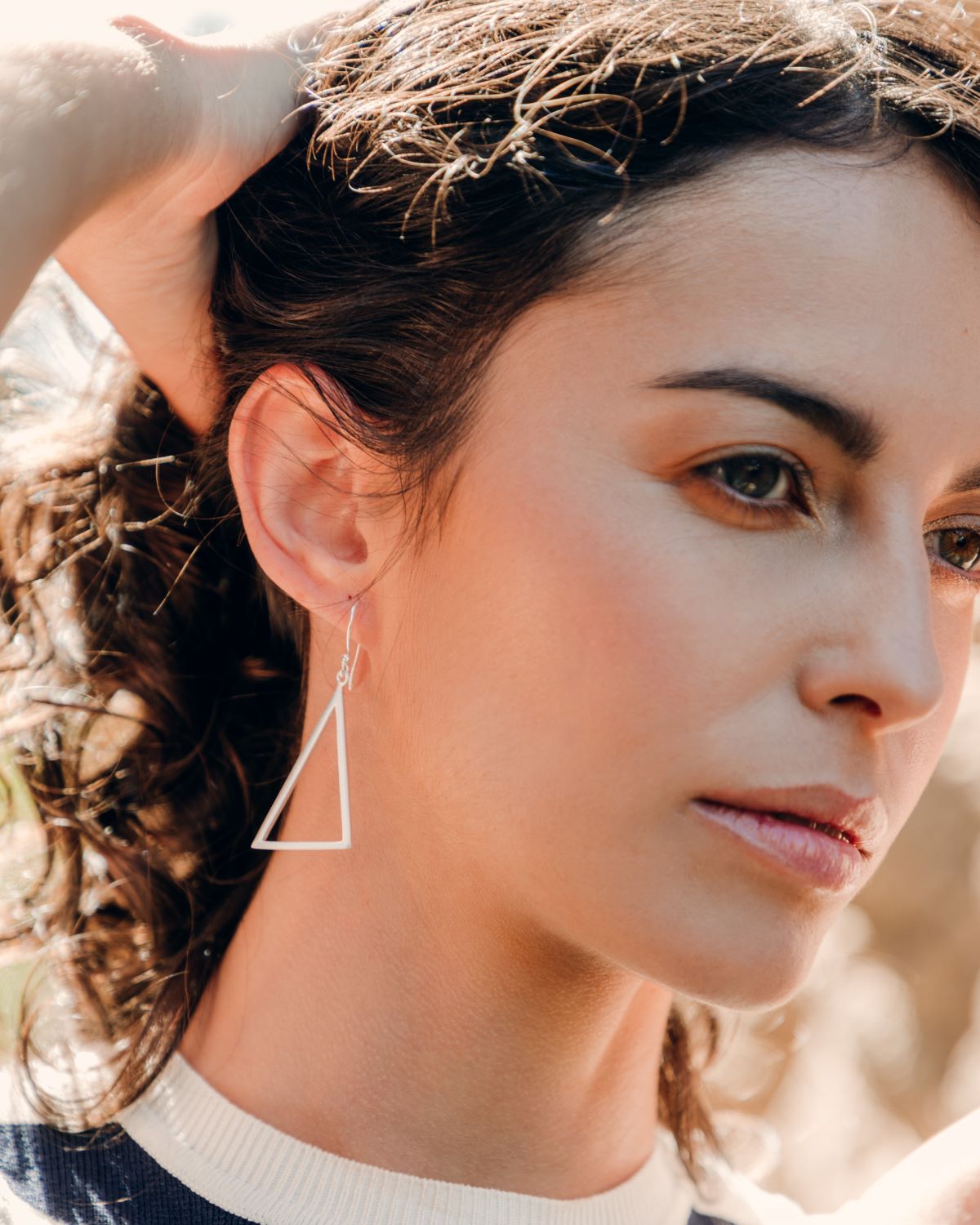 Pendientes triangulares plateados colgantes en la oreja de una modelo, en un plano muy cercano y con luz cálida.