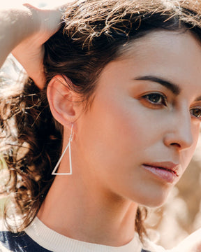 Pendientes triangulares plateados colgantes en la oreja de una modelo, en un plano muy cercano y con luz cálida.